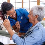 professional healthcare worker helping senior woman to apply lipstick at living room