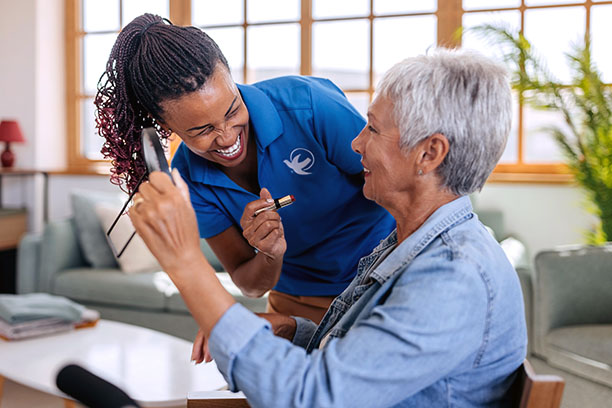 professional healthcare worker helping senior woman to apply lipstick at living room